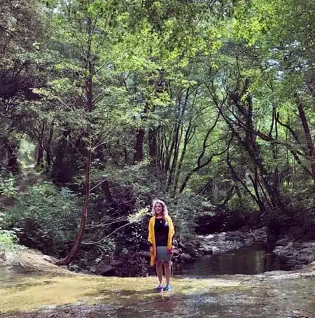 A woman standing in a forest stream surrounded by green.